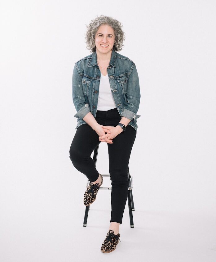 Leah Neaderthal, sales coach and business mentor, sitting on a stool, wearing a denim jacket and black pants, smiling confidently, promoting client acquisition and consulting success for women entrepreneurs.